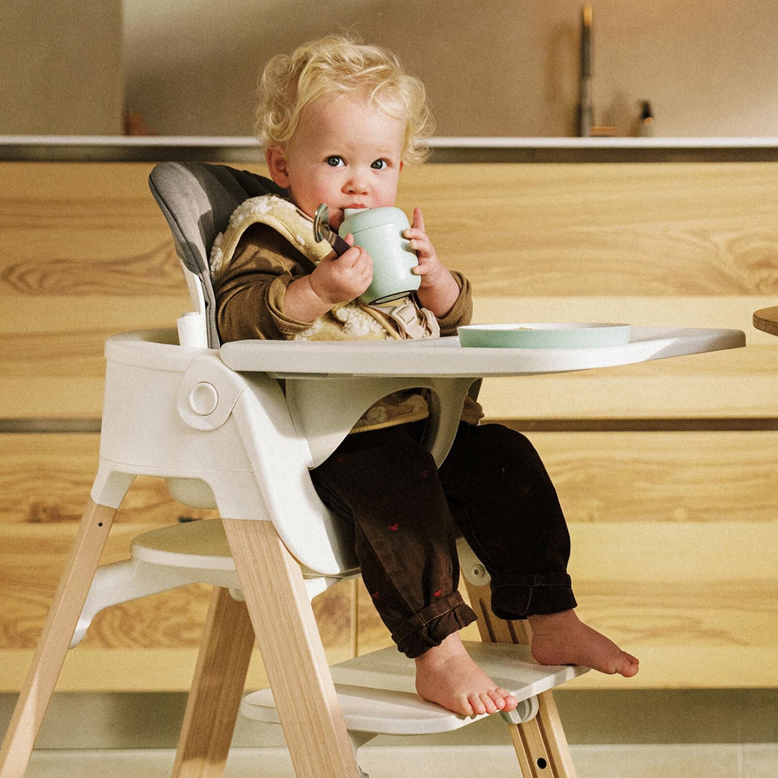 Een blonde peuter drinkt uit een beker in een witte kinderstoel met houten poten bij een aanrecht.