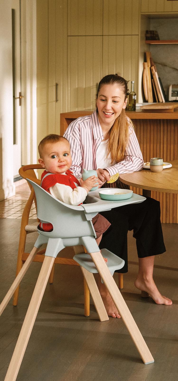 A smiling woman and a baby with messy face in a light blue high chair at a wooden table.