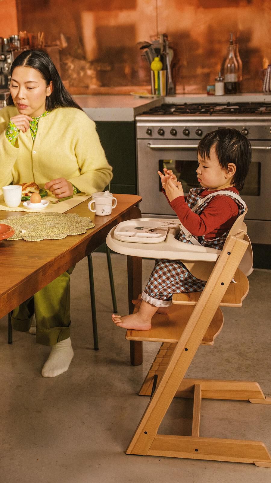 Eine Frau und ein Kind an einem Tisch. Das Kind sitzt in einem Tripp Trapp Hochstuhl und schaut auf seine Hände.