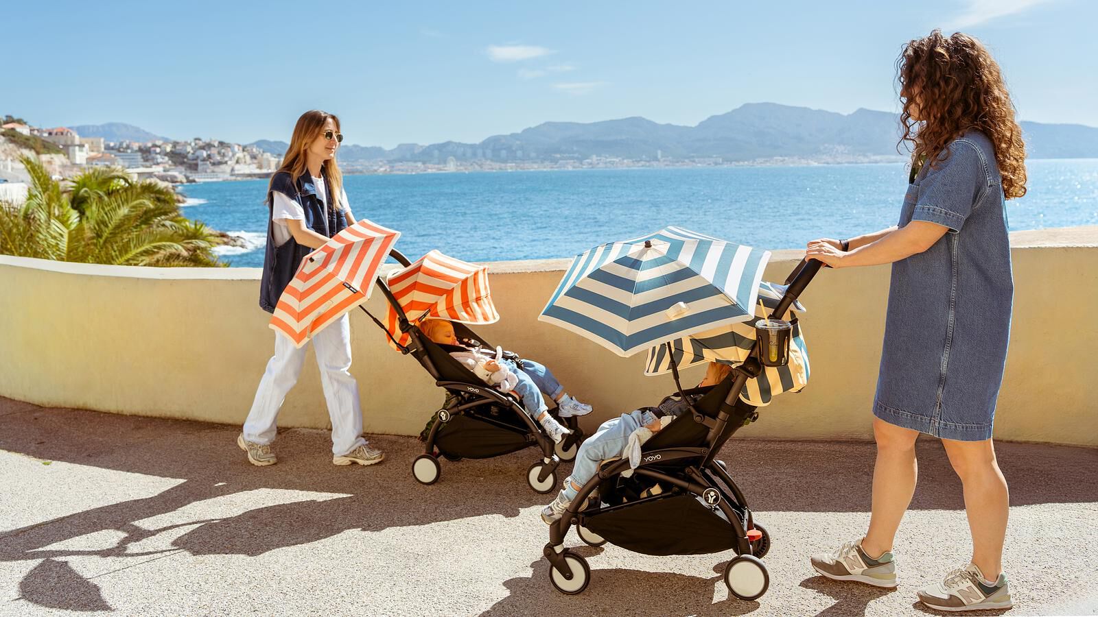 Two women push babies in YOYO strollers with umbrellas along a sunny coast.