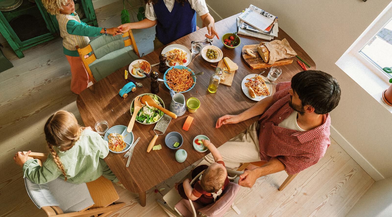 Family meal: parents and three children enjoy pasta at a wooden table, baby in a Tripp Trapp high chair.