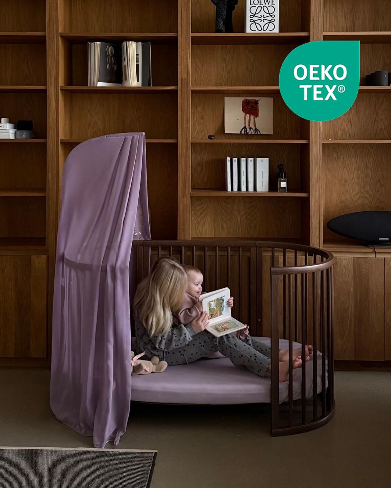 Un enfant plus âgé lit à un bébé dans un berceau rond en bois avec un baldaquin violet.