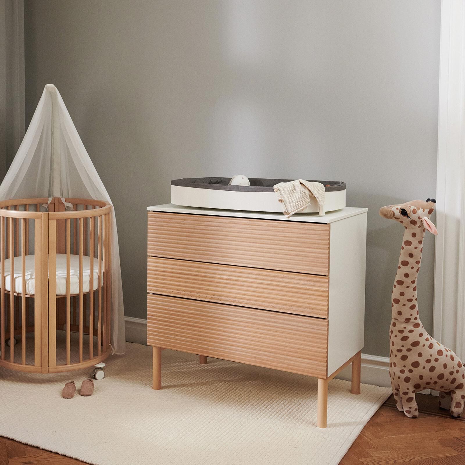 A cozy nursery with a wooden crib, dresser with changing pad, and a large stuffed giraffe.