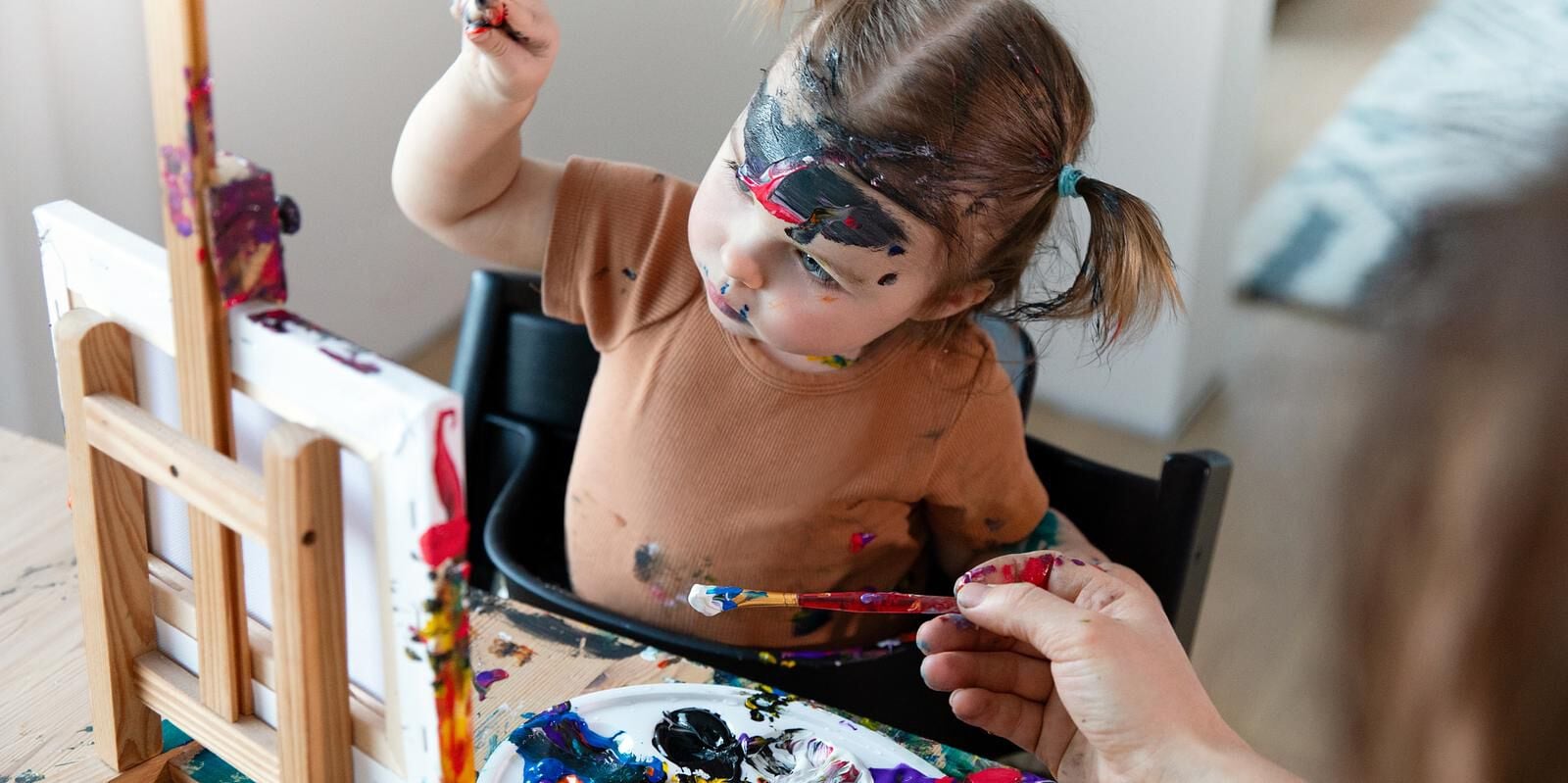 Een met verf besmeurd kind in een zwarte Tripp Trapp kinderstoel reikt naar een penseel.