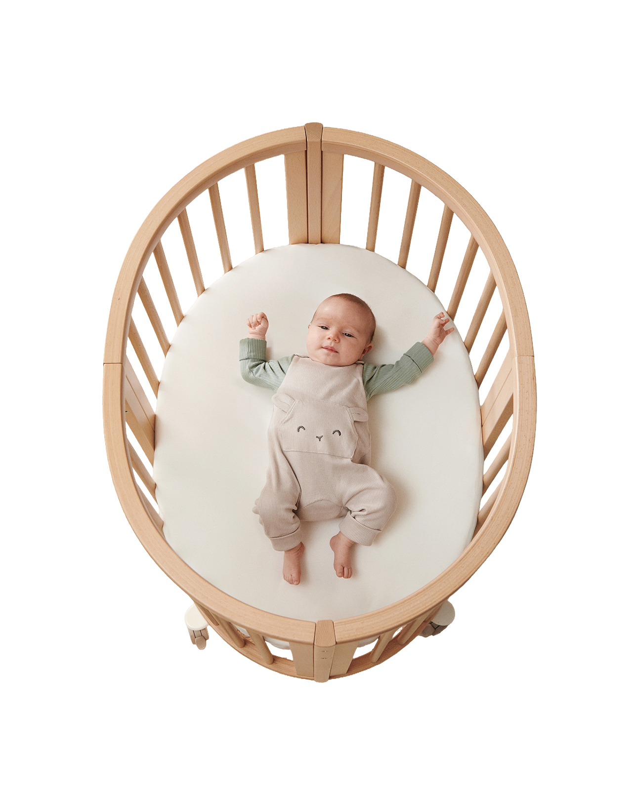 A baby in bear overalls lies on a white mattress in an oval wooden crib.