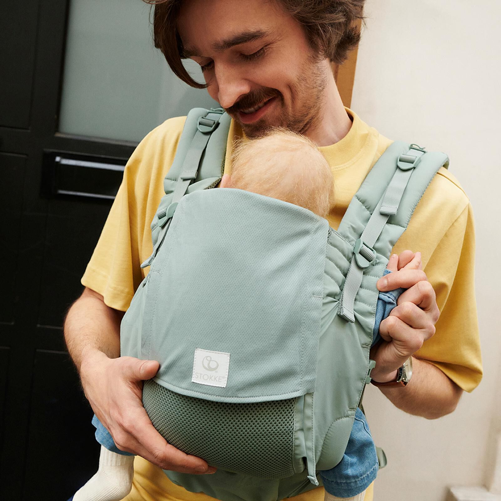 Un homme souriant porte un bébé dans un porte-bébé vert.