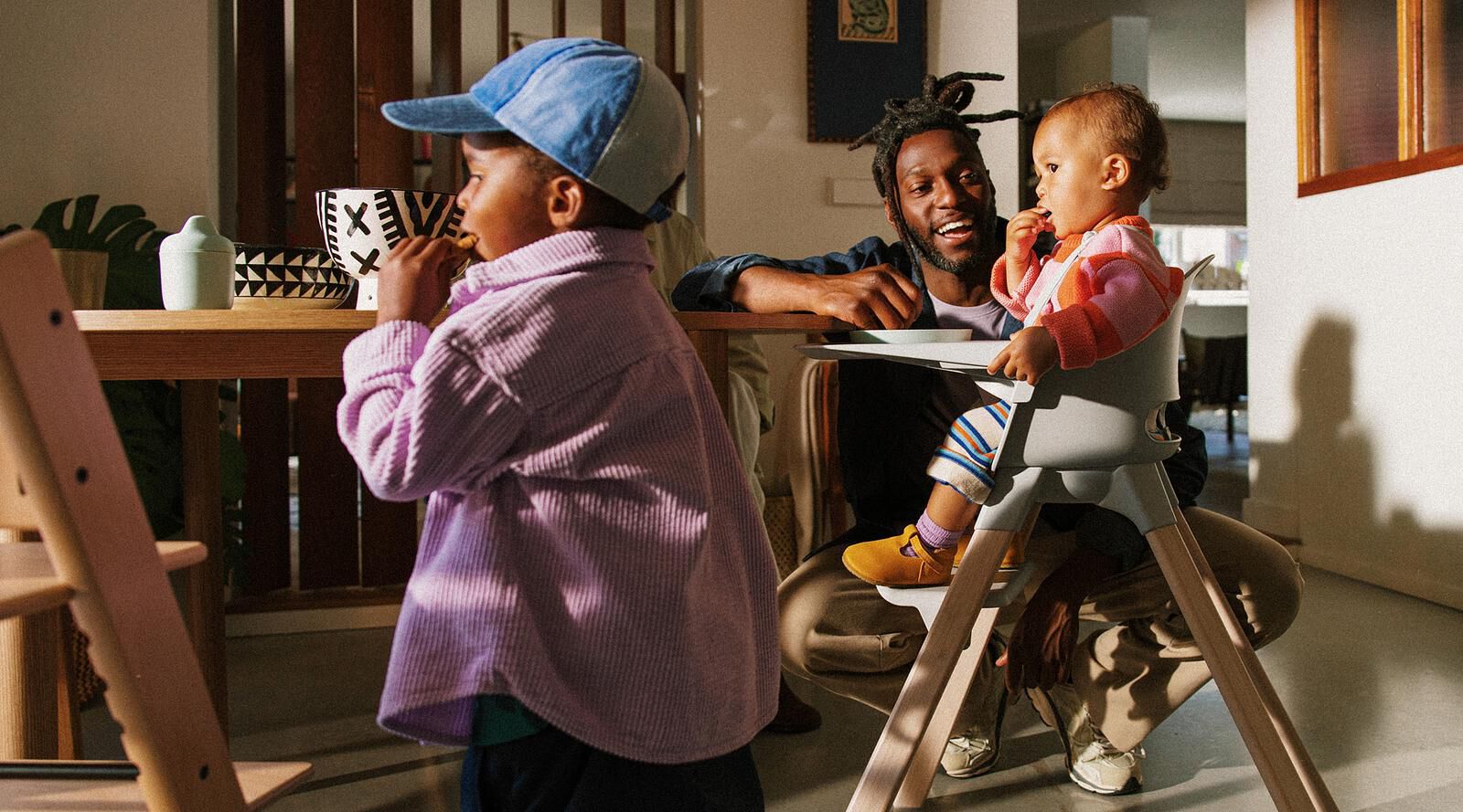 A father smiles at his baby eating in a Tripp Trapp high chair, while another child stands nearby.
