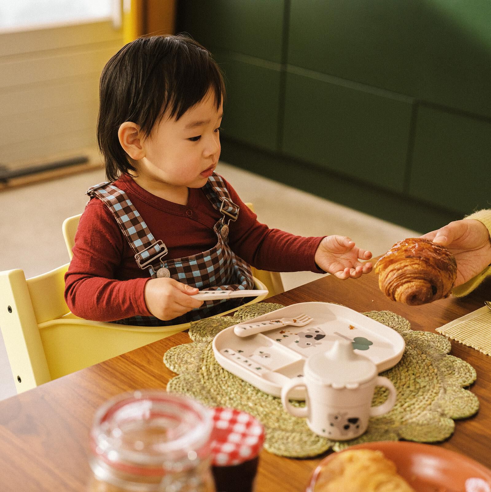 Småbarn i en gul Tripp Trapp barnestol strekker seg etter en croissant fra en voksenhånd.