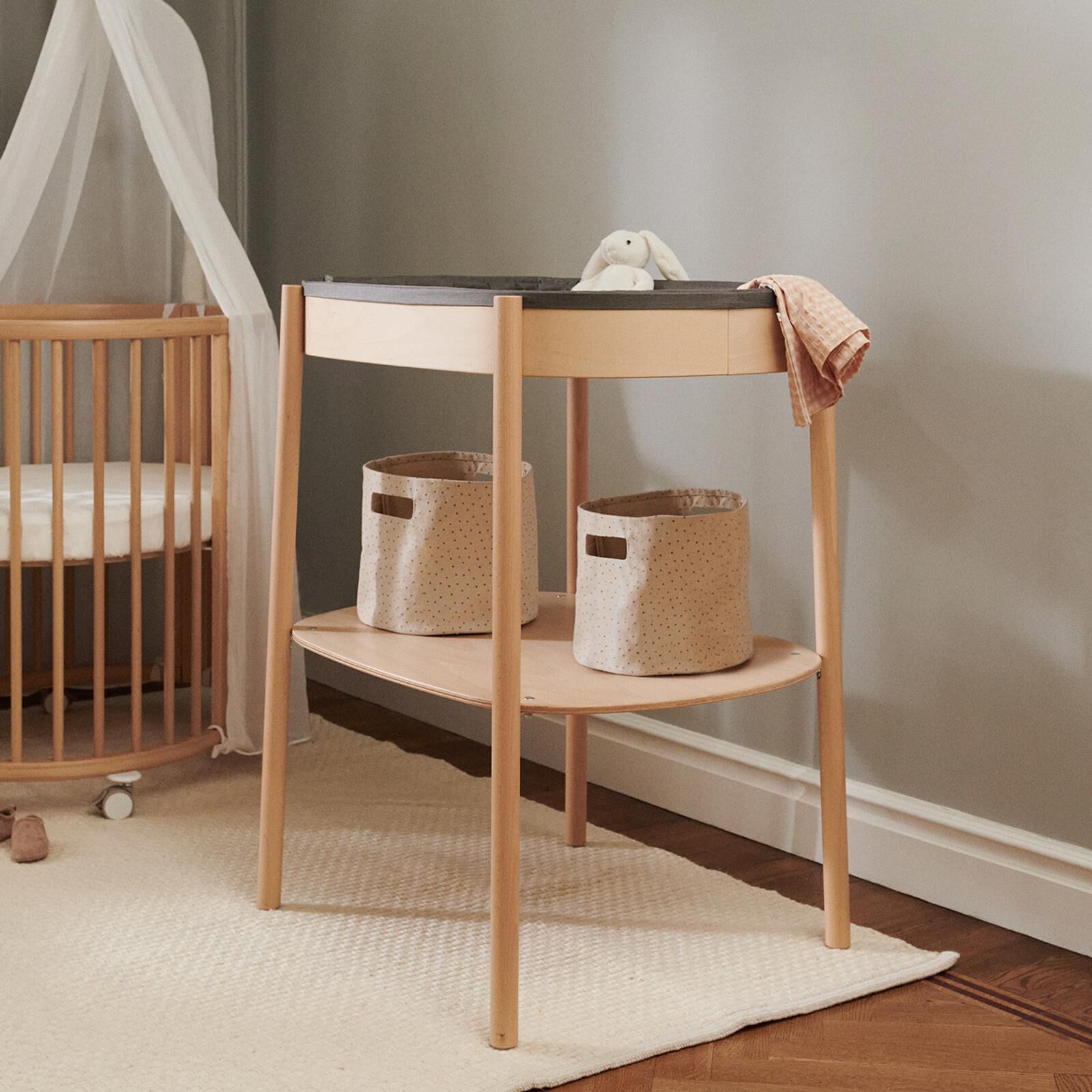 Ein Wickeltisch aus Holz mit einem Hasenspielzeug und Tuch, zwei Aufbewahrungskörben und einem Kinderbett.
