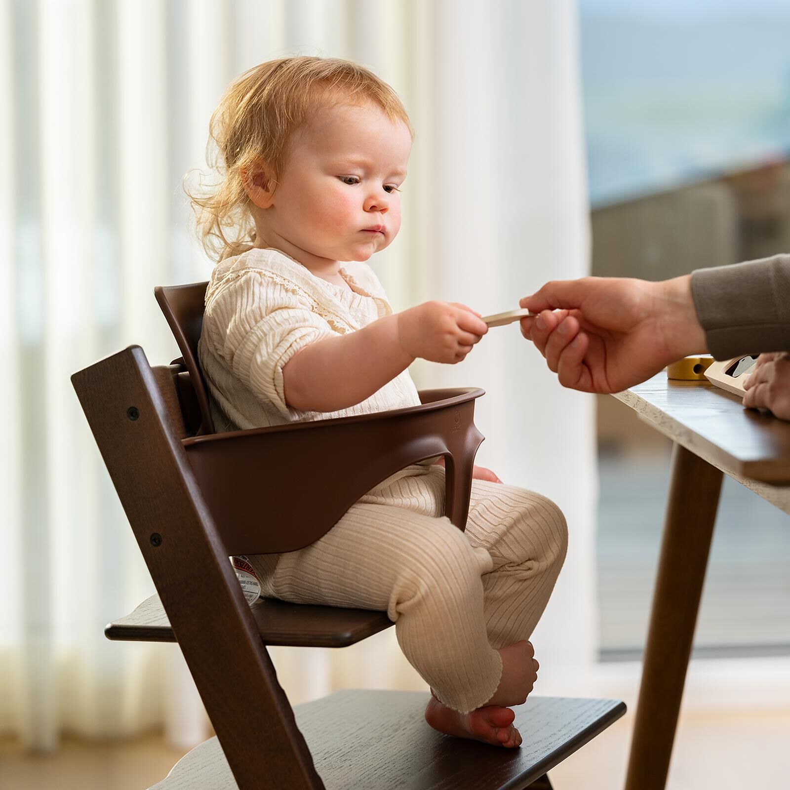 A baby in a dark brown Tripp Trapp high chair reaches for a wooden toy from an adult.