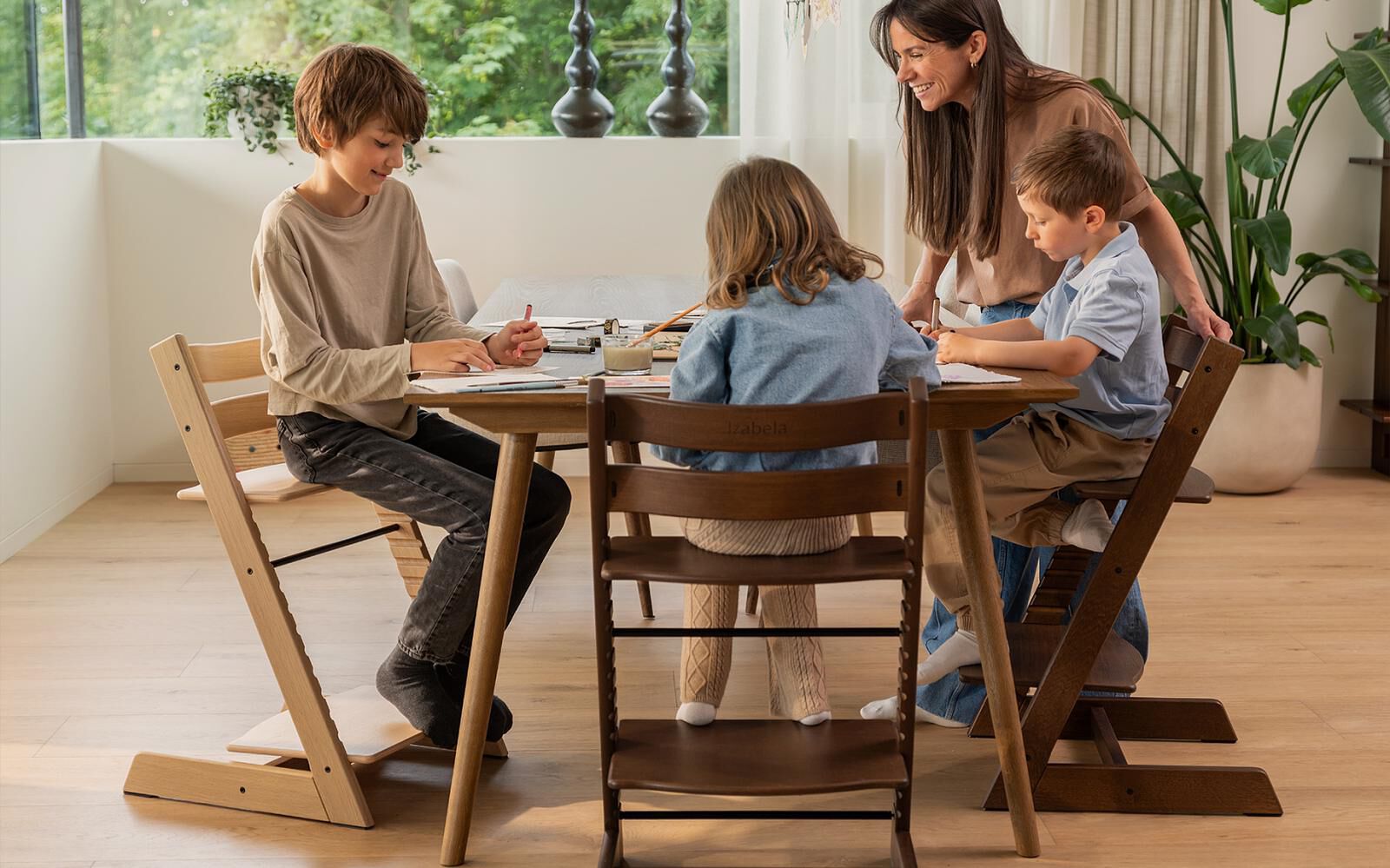 Een vrouw glimlacht terwijl ze drie kinderen aan een tafel ziet tekenen, allemaal zittend in Tripp Trapp stoelen.