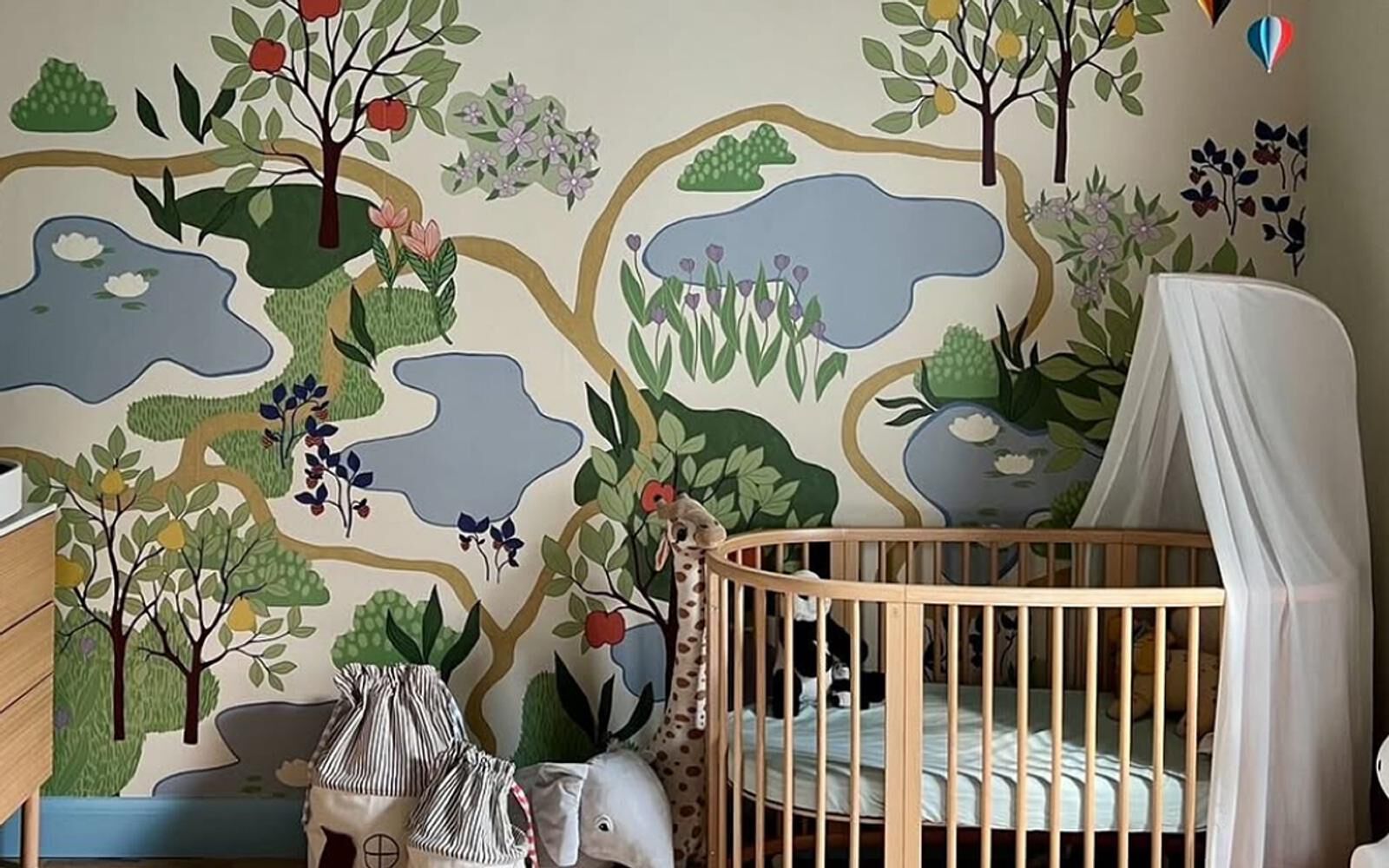 Une chambre d'enfant douillette avec une fresque murale sur le thème de la nature, comprenant un berceau rond en bois, un baldaquin blanc et des animaux en peluche.