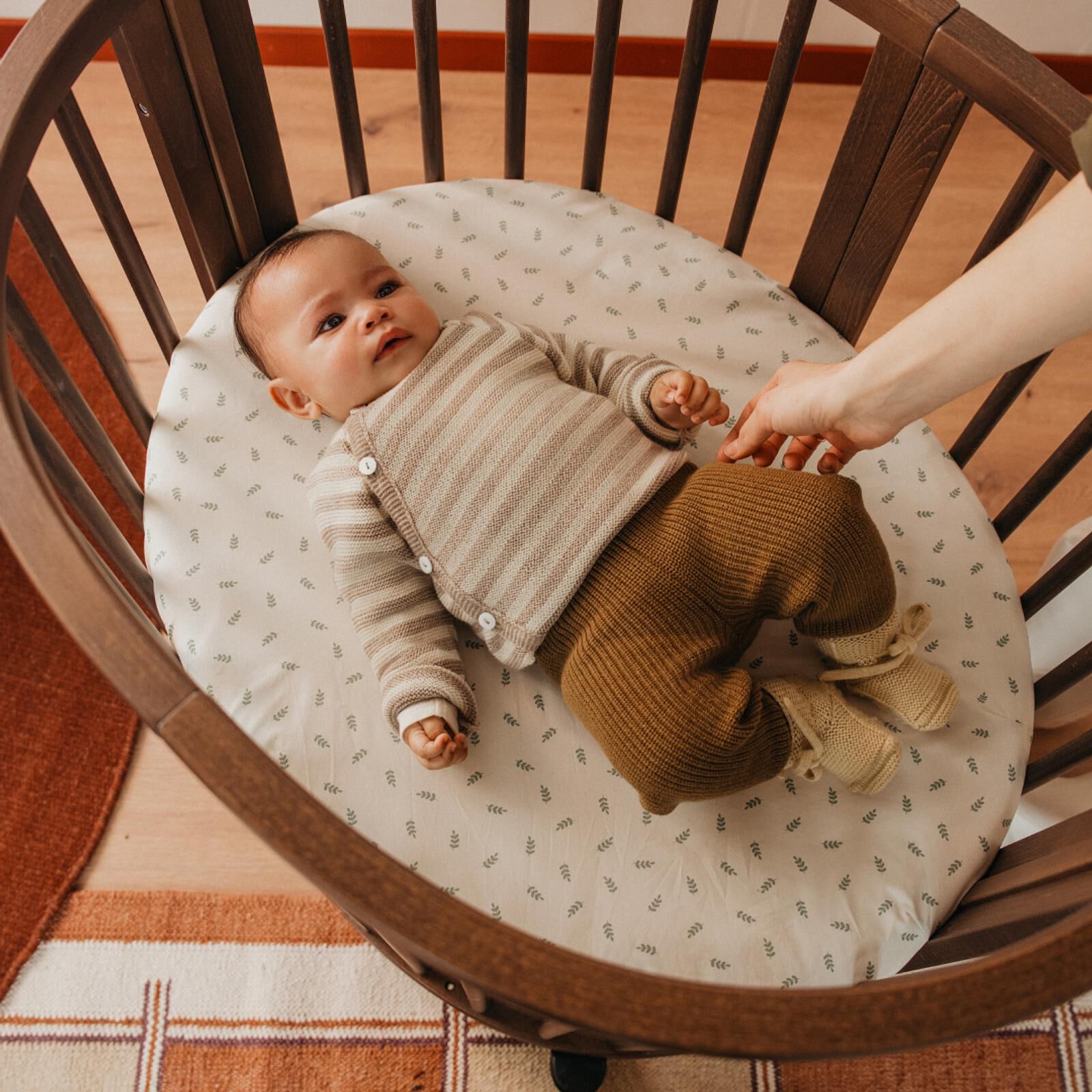 Baby ligt in een ronde houten wieg, draagt een gestreepte trui, terwijl een hand zachtjes hun been aanraakt.