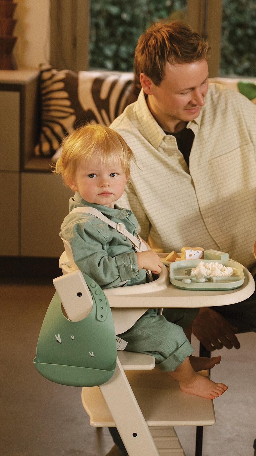 Blonde peuter in een Tripp Trapp kinderstoel met een groen slabbetje en dienblad kijkt naar de camera terwijl een volwassene glimlacht.