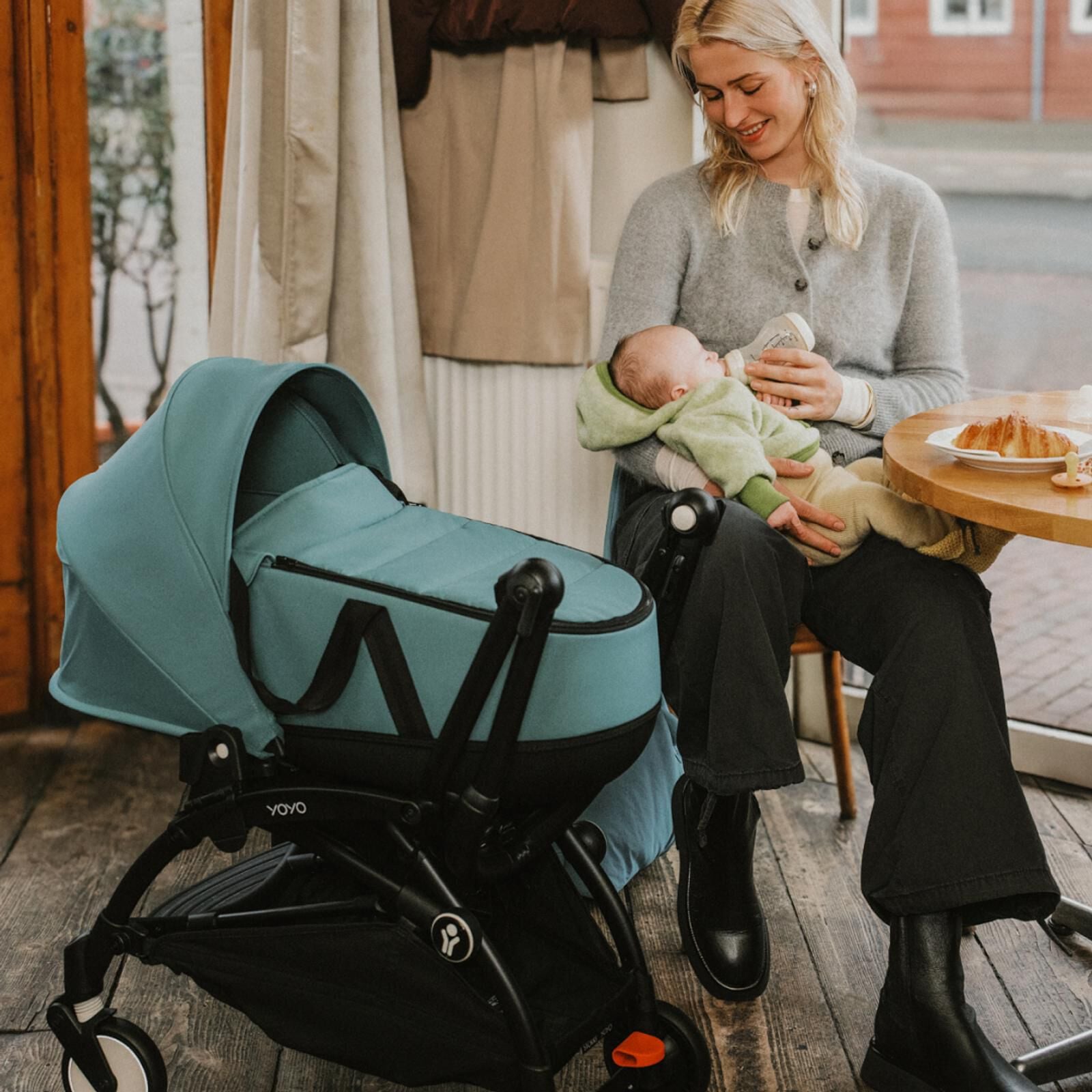 Een vrouw voedt een baby in een café, met een STOKKE YOYO kinderwagenwiegje naast hen.