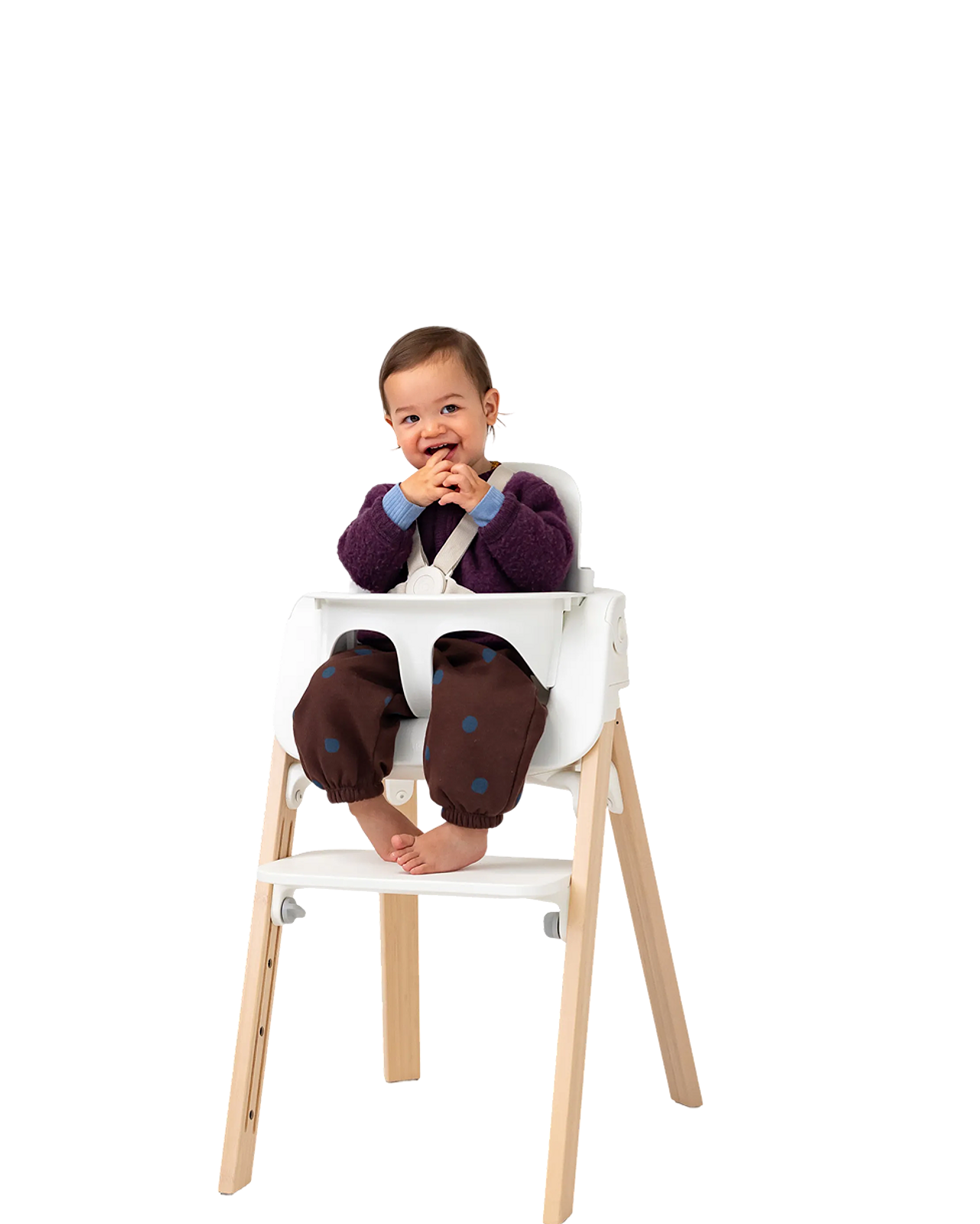 A happy baby sits in a white Tripp Trapp high chair with natural wood legs, smiling at the camera.