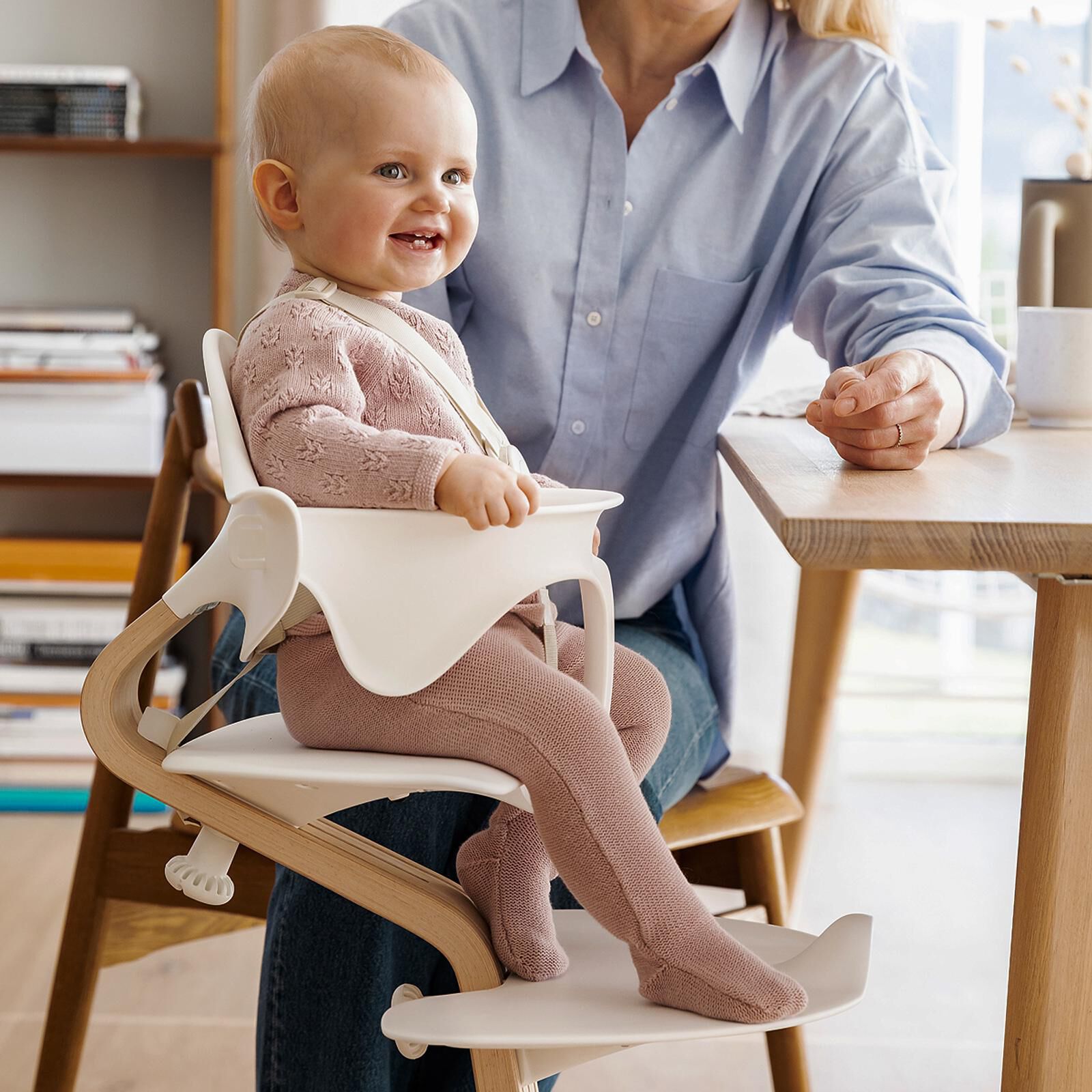 Lachende baby in een Tripp Trapp kinderstoel met Baby Set, naast een volwassene aan een houten tafel.