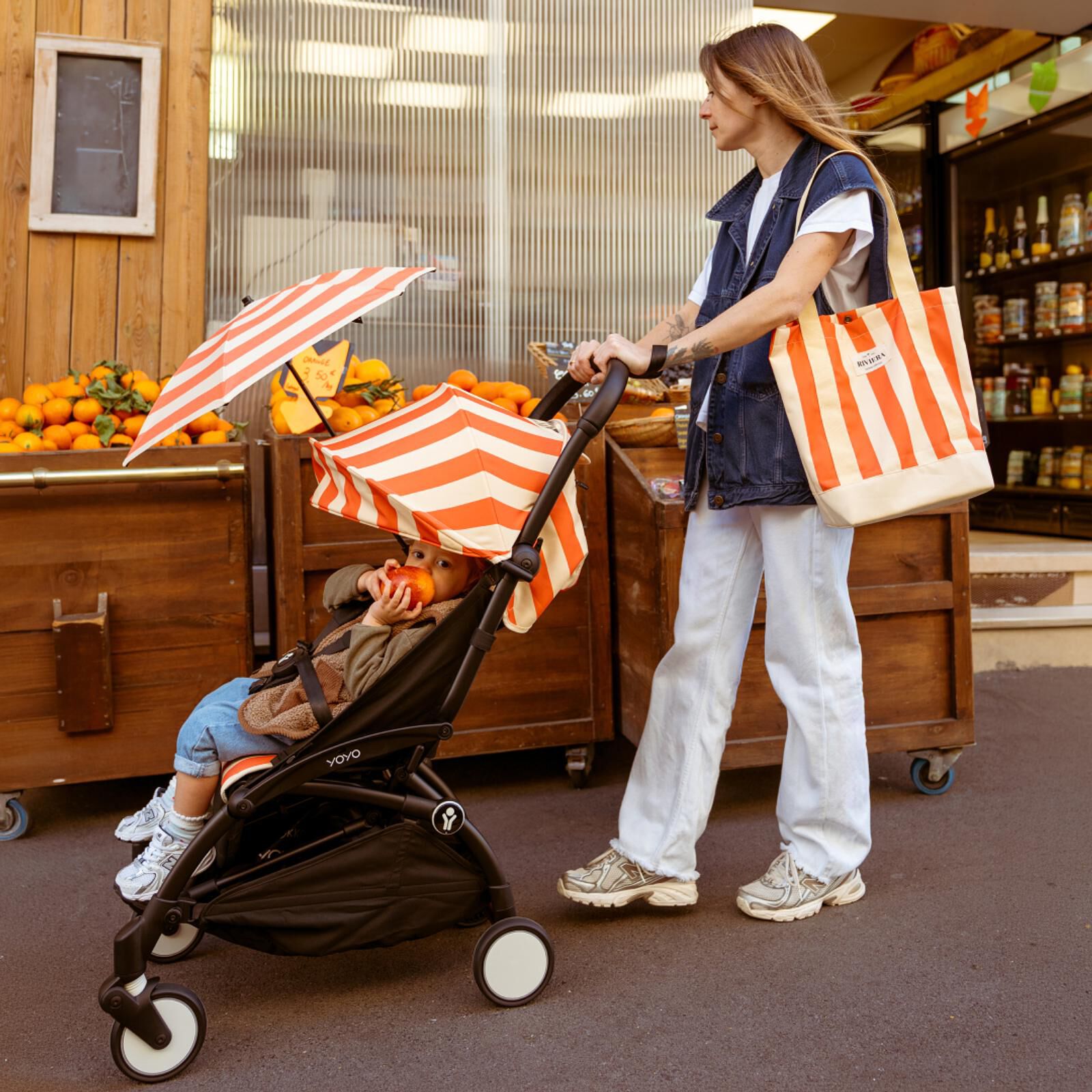 Woman pushes a child eating an apple in a striped STOKKE YOYO stroller at a fruit stand.