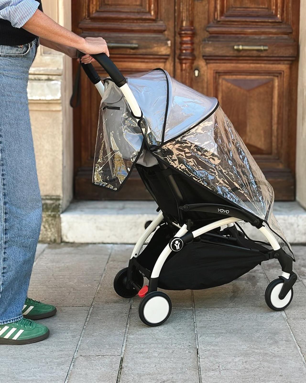Eine Person schiebt einen YOYO Kinderwagen mit Regenschutz vor Holztüren.
