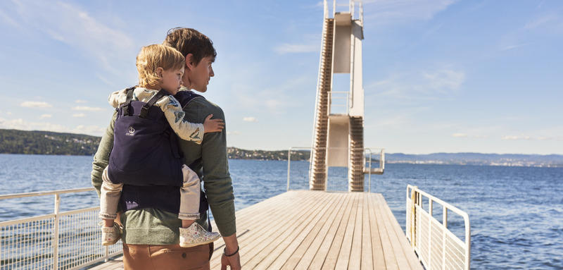 Dad carrys child on back by ocean with Stokke MyCarrier