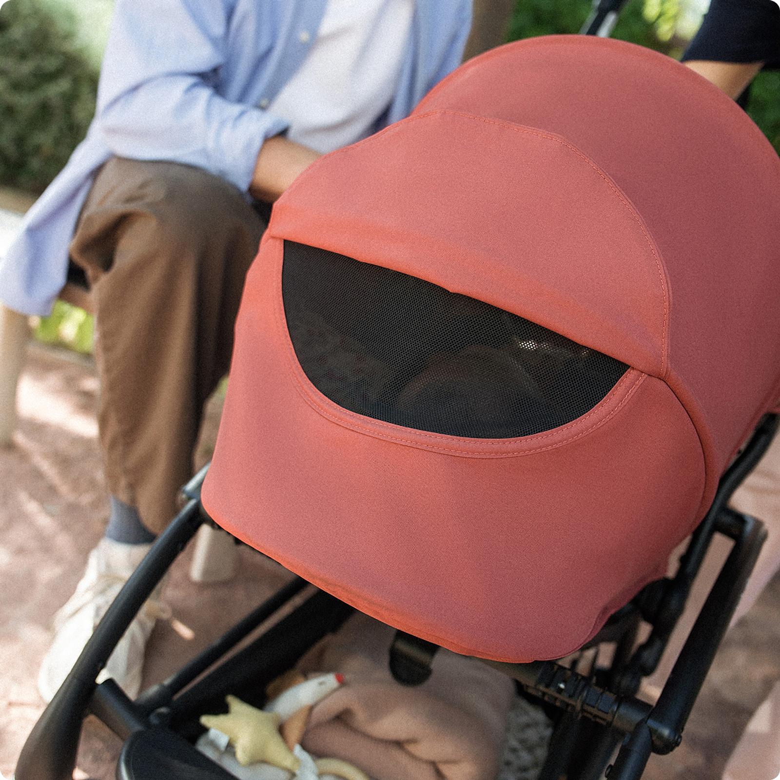 A reddish-orange STOKKE YOYO stroller with a mesh peek-a-boo window, outdoors.