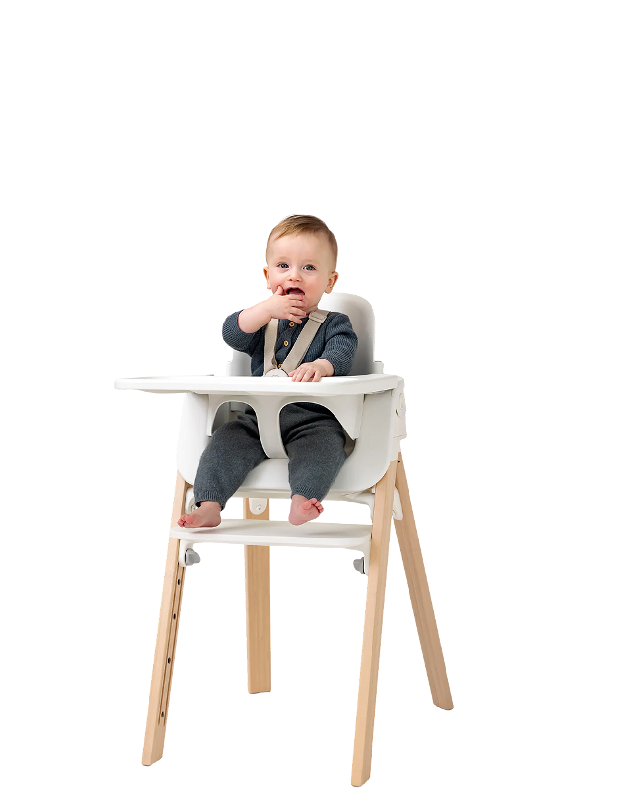 A baby sits in a white Tripp Trapp high chair, hand near mouth.
