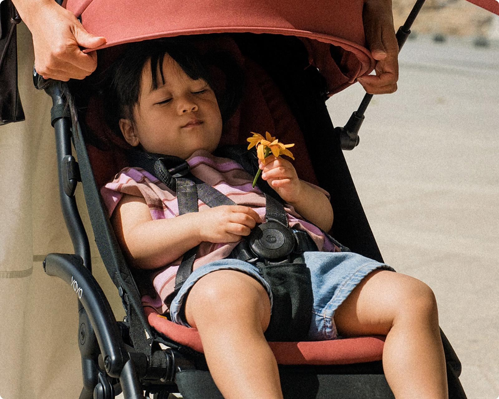 A sleeping child holds a yellow flower in a STOKKE YOYO stroller with a parent's hands.