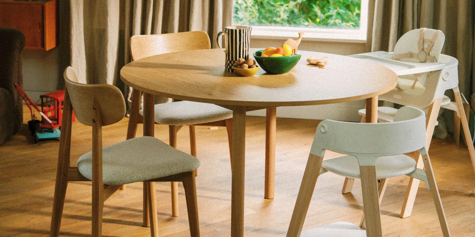 A round wooden dining table surrounded by chairs and a white high chair, with fruit and nuts on the table.