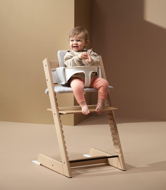 Baby sitting in natural tripp trapp high chair with white baby set and grey cushion.