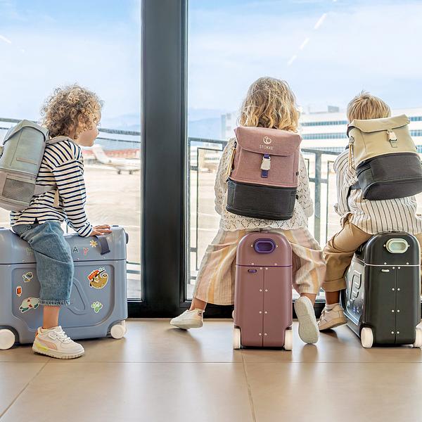 Three children are looking out an airport window while sitting on JetKids luggage.​