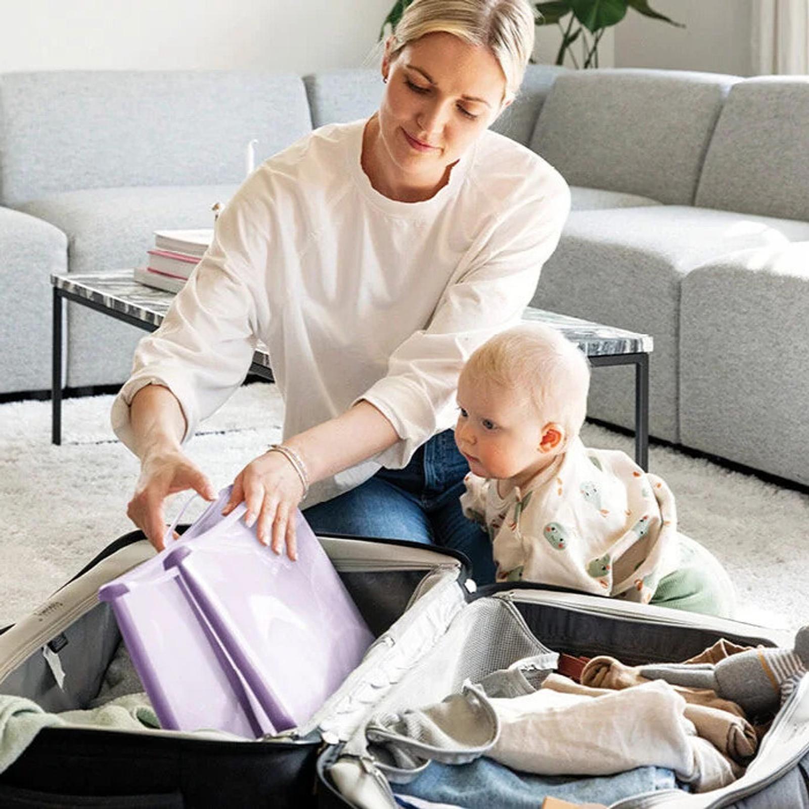 A mother packs a suitcase with a purple item, her baby watches.