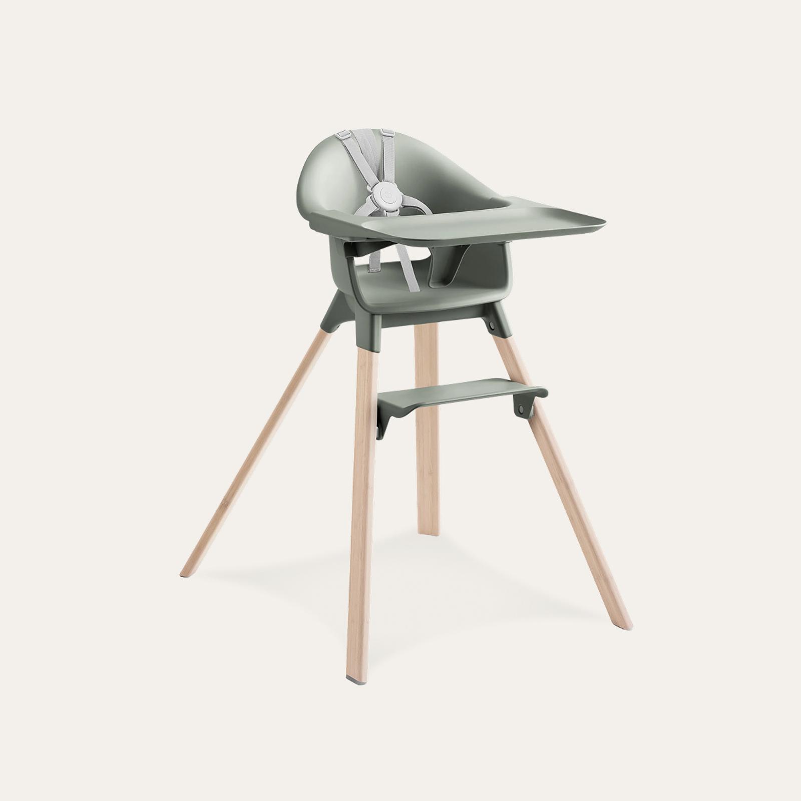 A modern high chair with sage green seat, tray, and footrest, on light wooden legs with a grey harness.