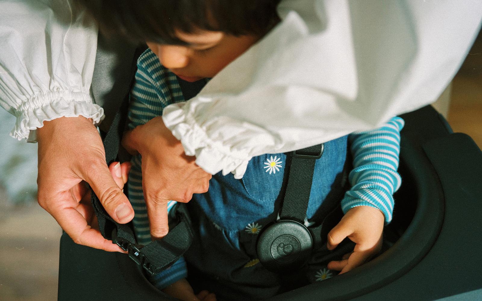 Adult buckling a child into a STOKKE YOYO high chair. Child wears a daisy overall.