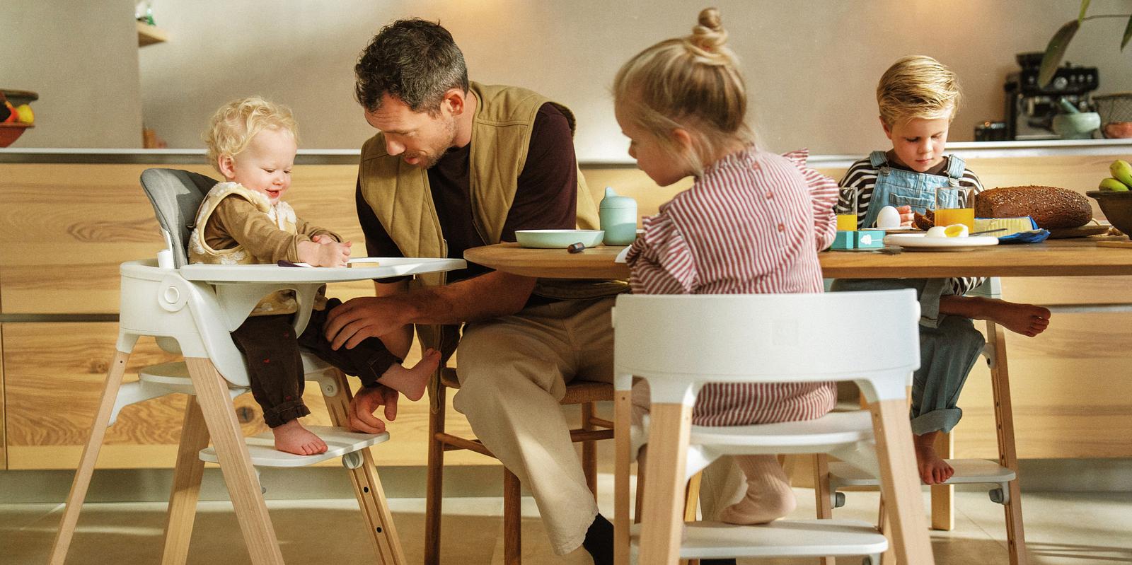Father and three children eating. One child in a Tripp Trapp high chair, two in Tripp Trapp chairs.