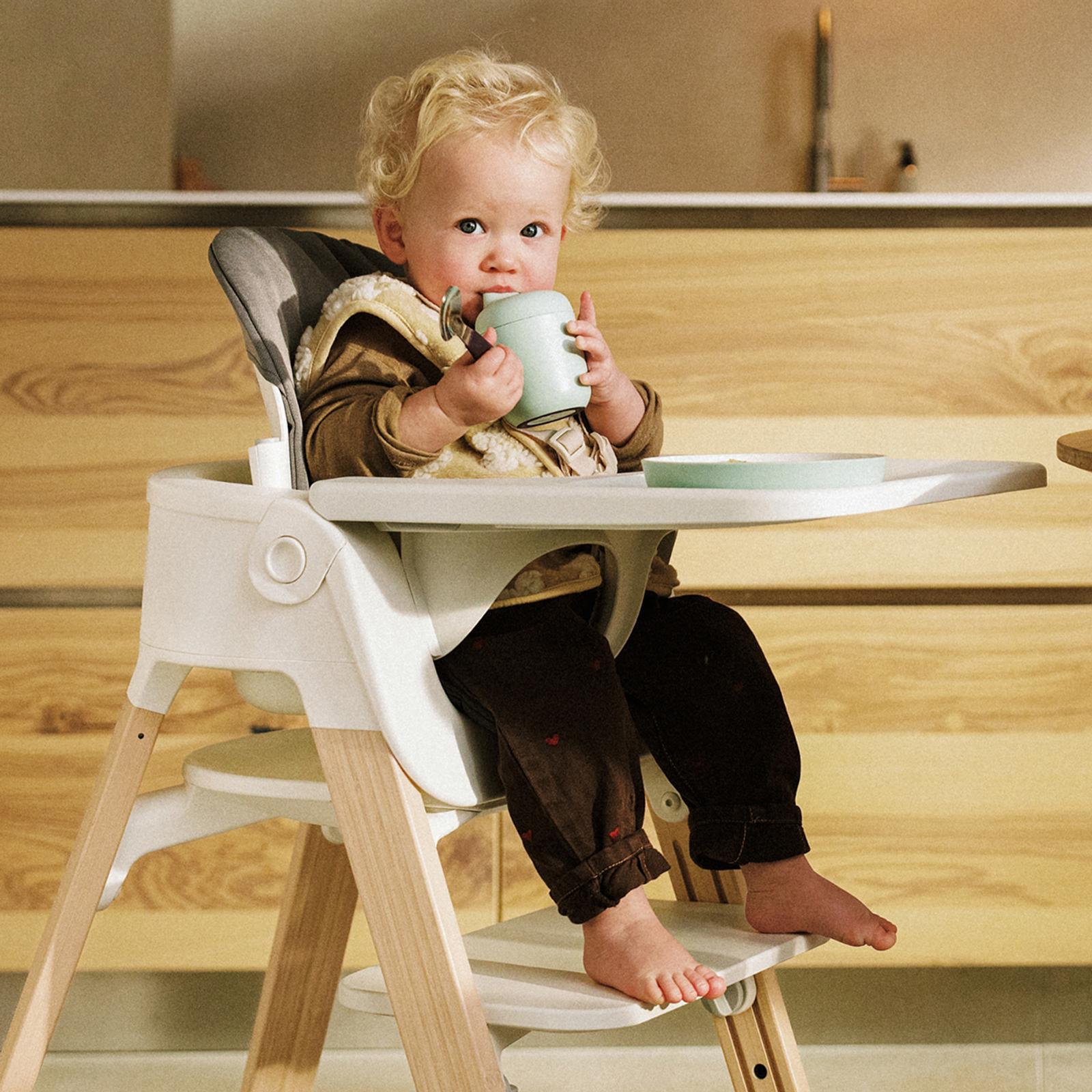 Blonde toddler drinks from a light green cup in a white high chair with wooden legs.
