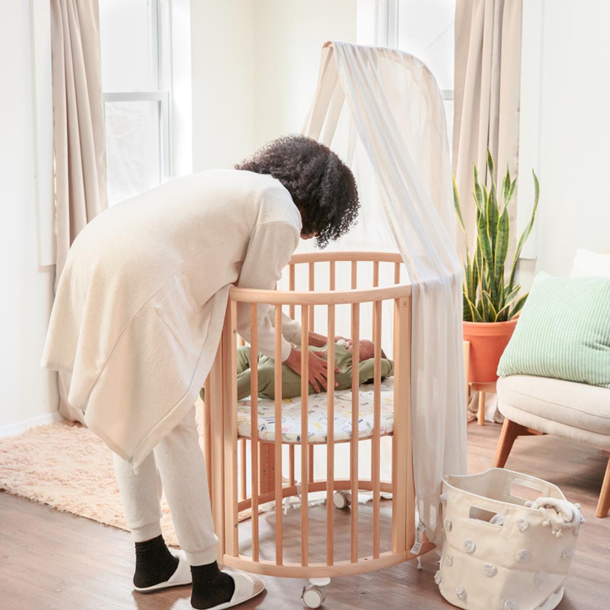 A mother putting her newborn baby in the sleepi mini crib in color natural. A great newborn baby gift.