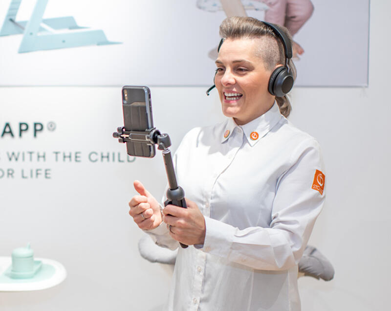 Woman holding a phone on a selfie stick wearing headphones explaining Stokke product through our live shopping online feature.