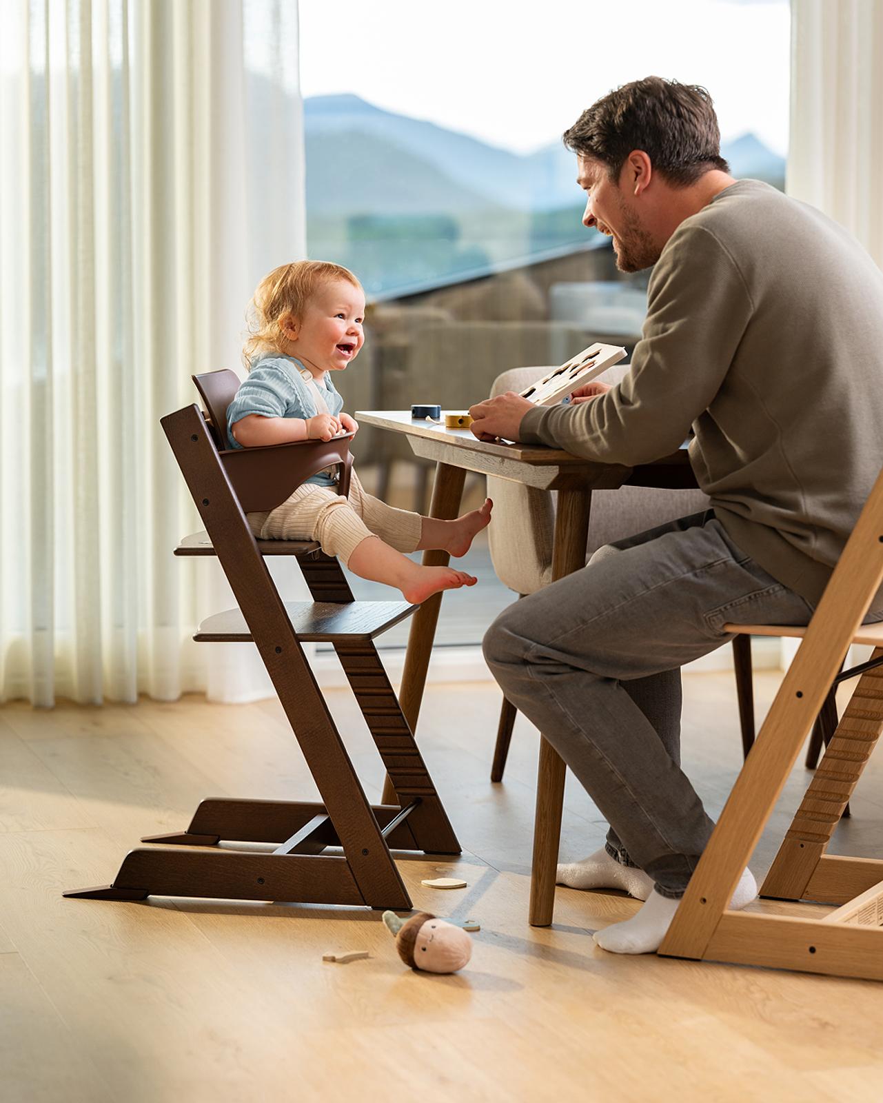 A baby laughs in a brown Tripp Trapp high chair as a man plays a puzzle.