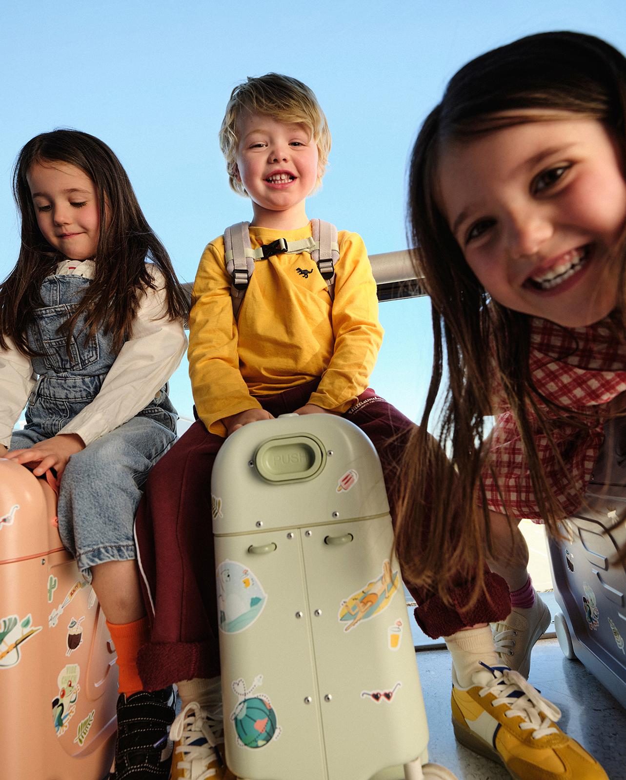 Three smiling children sit on colorful, sticker-covered ride-on suitcases under a blue sky.
