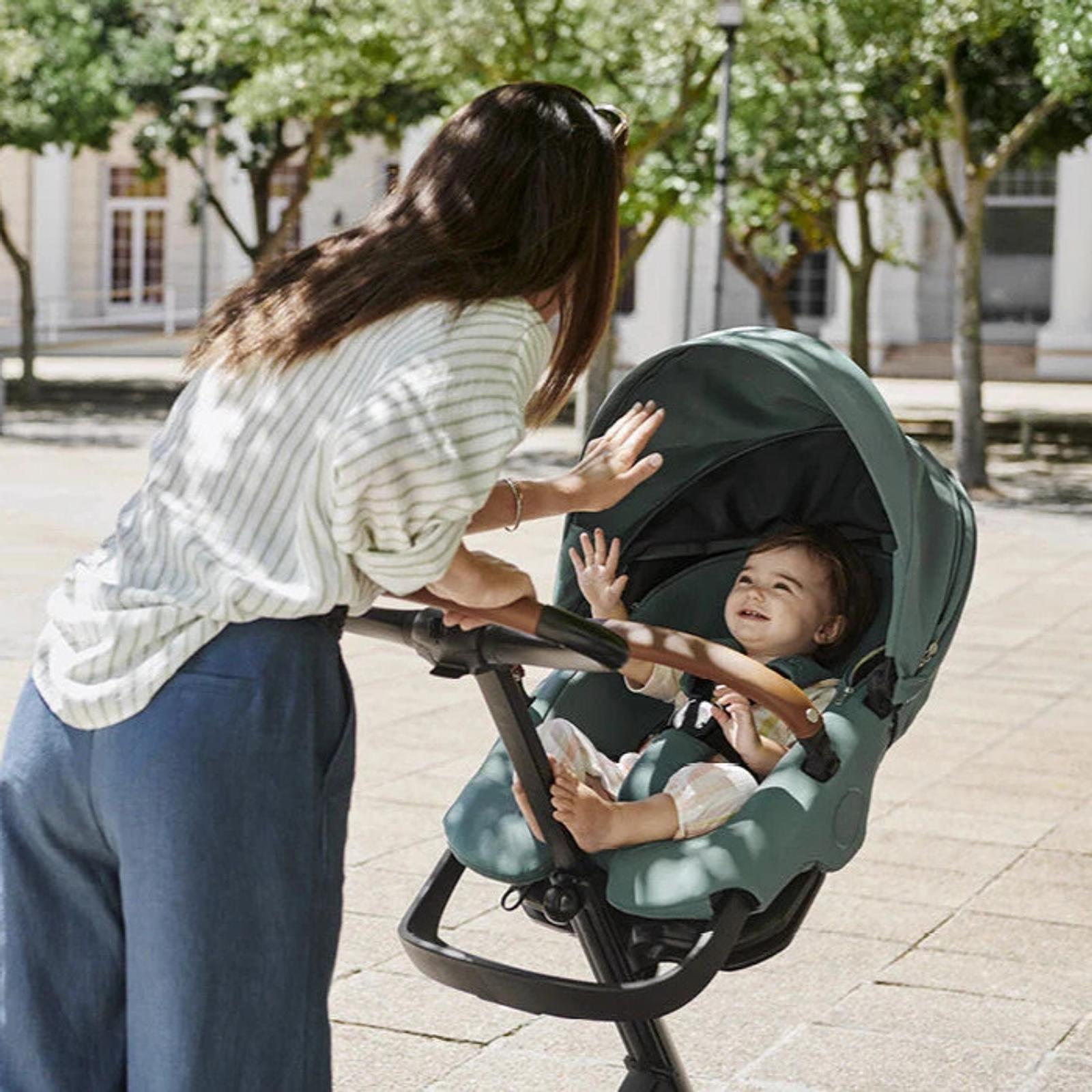 女性が屋外で緑色のSTOKKE YOYOベビーカーに乗ったご機嫌な赤ちゃんと交流している。
