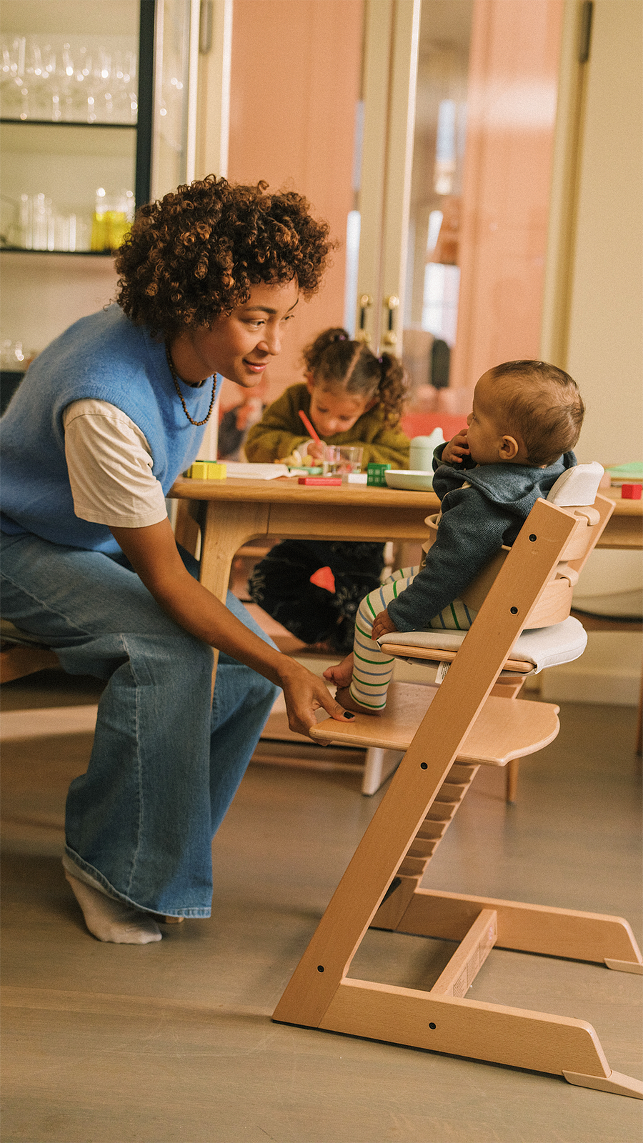 A woman interacts with a baby in a Tripp Trapp high chair, while a toddler plays at the table.