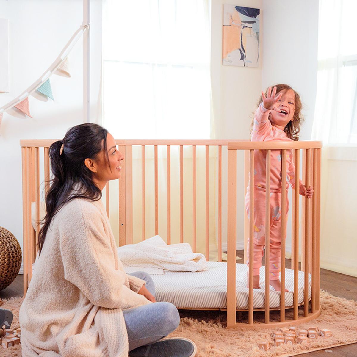 Young girl standing in her sleepi bed crib color natural in the nursery with her mom.