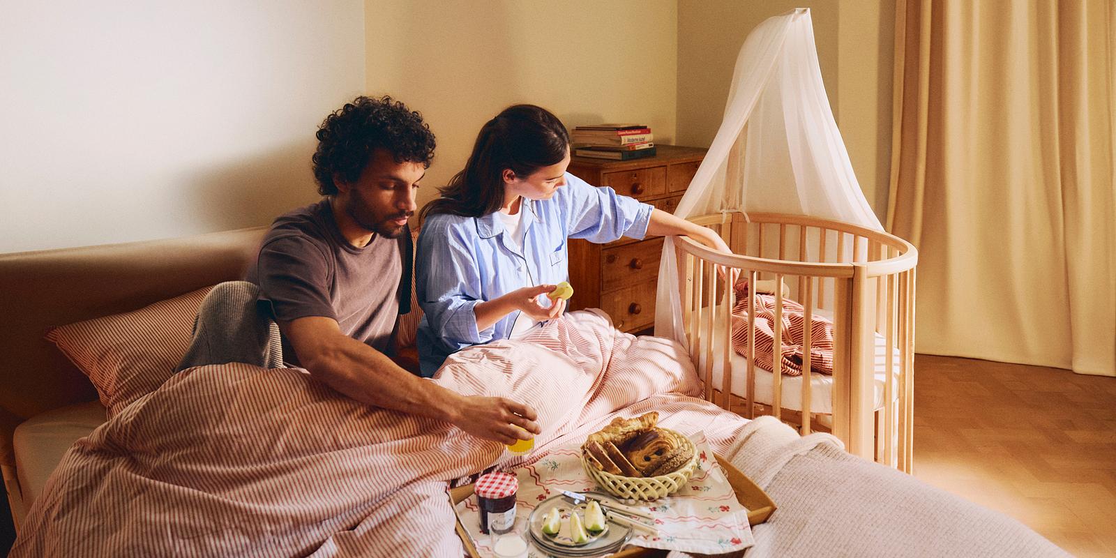 夫婦がベッドで朝食をとり、女性がSTOKKE Sleepiベビーベッドに手を伸ばしています。