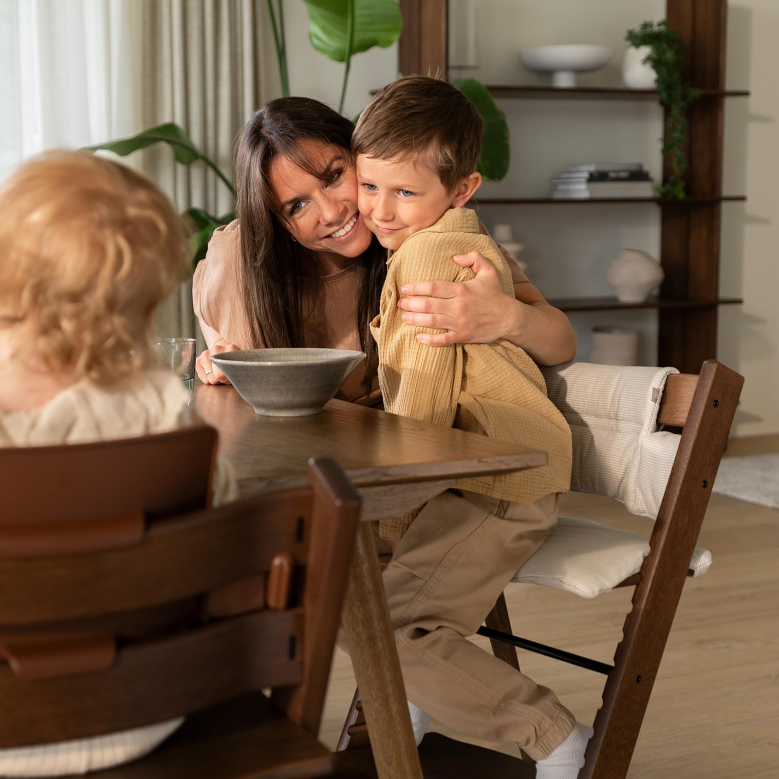 A happy mother hugs her son sitting in a Tripp Trapp chair at a dining table.