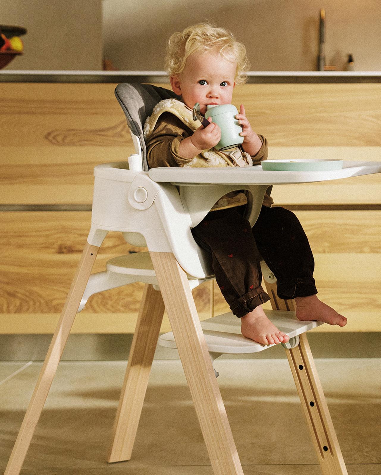 A blonde baby drinks from a green sippy cup in a white and wood high chair in a kitchen.