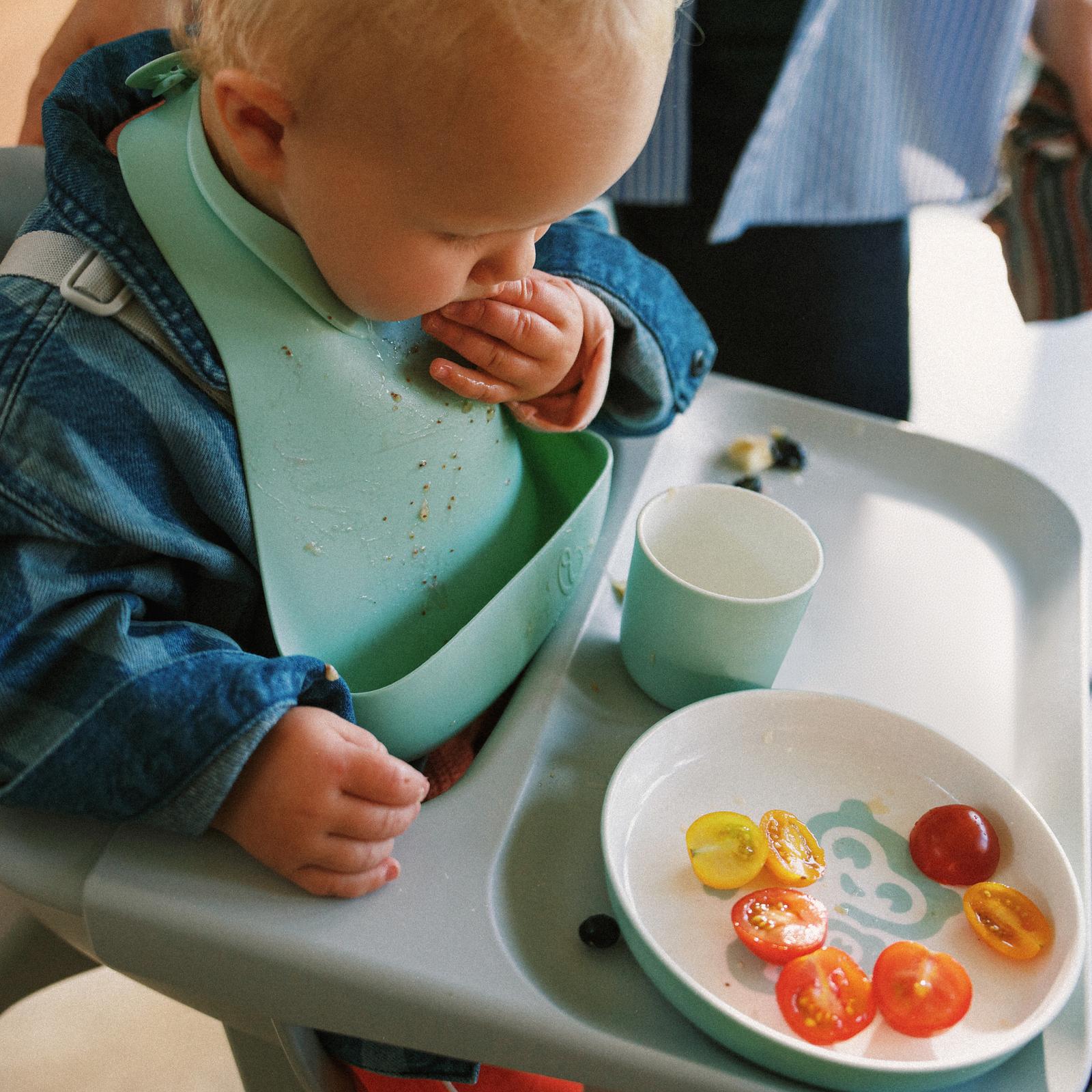 Ein Baby in einer Jeansjacke und einem grünen Lätzchen isst Kirschtomaten von einem Teller in einem grauen Hochstuhl.