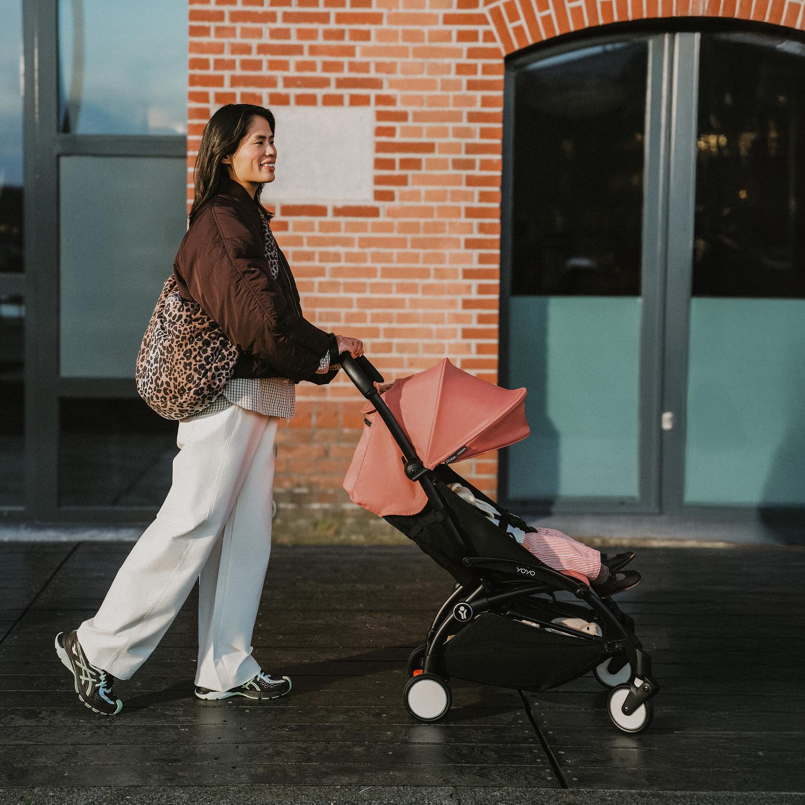 晴れた日、笑顔の女性がサンゴ色のキャノピーが付いたSTOKKE YOYOベビーカーを押しています。