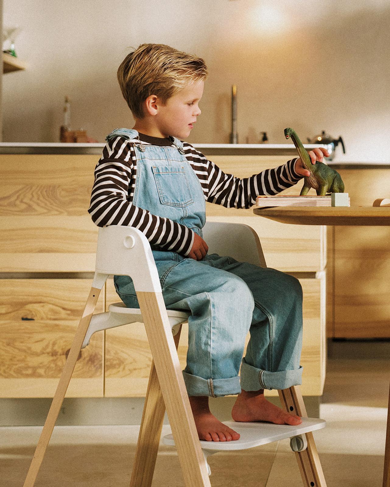 A boy in a high chair plays with a dinosaur toy at a wooden table.