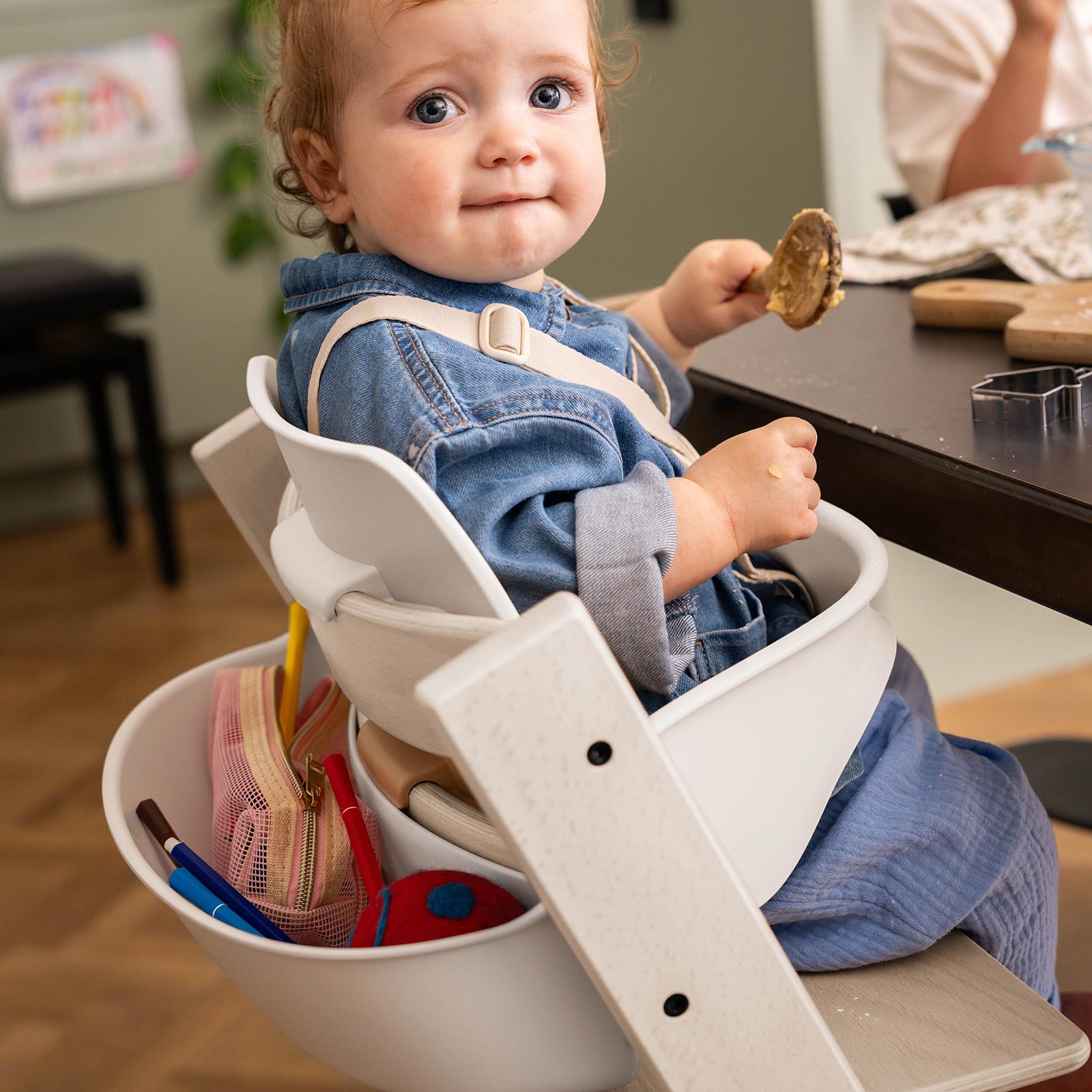 A baby sits in a white Tripp Trapp high chair, holding a spoon, with art supplies on the side.