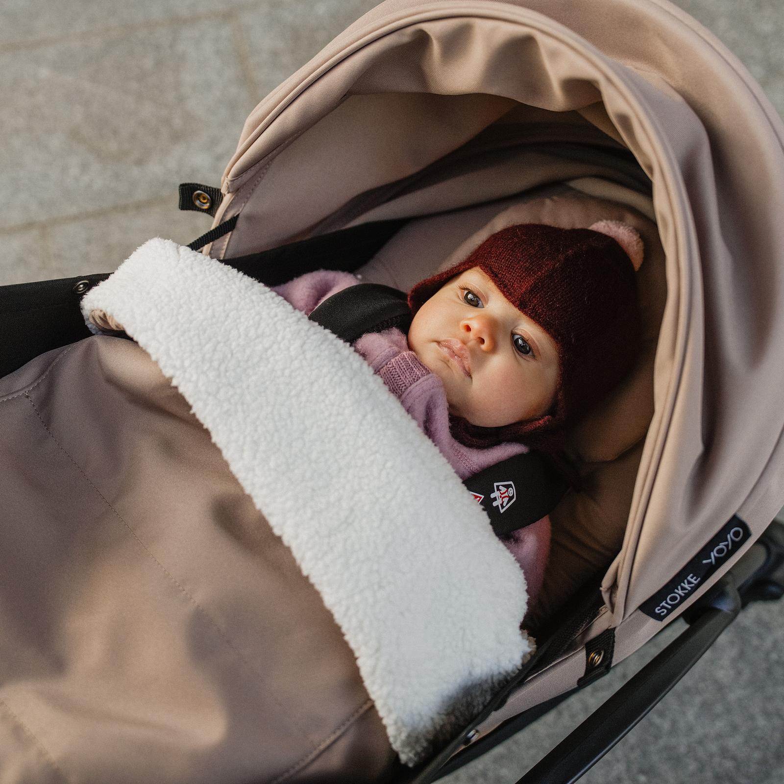 Ein Baby mit burgunderfarbenem Hut und rosa Kleidung, eingewickelt in eine weiße Decke, in einem beigefarbenen STOKKE YOYO Kinderwagen.